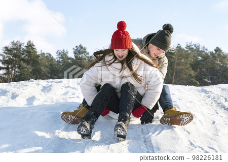 Young guy and a girl slide down the snow slide against the background of the forest and the blue sky. Young guy and a girl slide down the snow slide against the background of the forest and the blue sky. 98226181