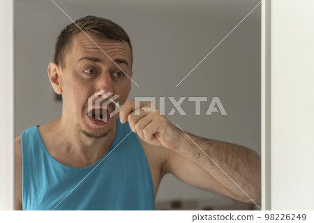 Unhappy young man in blue T-shirt removing unwanted hair in nose standing in front of mirror. Morning routine Unhappy young man in blue T-shirt removing unwanted hair in nose standing in front of mirror. Morning routine 98226249