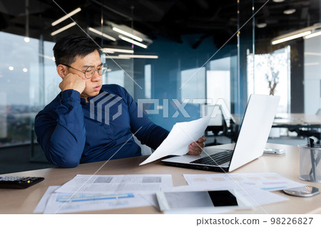 Tired young Asian man, accountant, businessman, freelancer. He sits in the office at a table with a laptop, looks through documents, holds his head in his hands, sighs a hard and boring day. 98226827