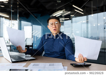 Problems at work. Worried young Asian man, businessman, freelancer. He sits in the office at a table with a laptop, holds documents in his hands. Spreads his hands, looks at the camera confused. 98226828