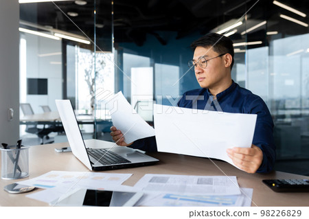 A serious young man, an Asian businessman, works in the office at the table, holds and looks at documents, graphs, plans, bills. 98226829