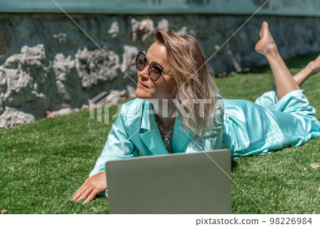 a young beautiful woman with blond curly hair in glasses and a blue dress sits on the grass in nature and uses a laptop, 98226984