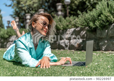 a young beautiful woman with blond curly hair in glasses and a blue dress sits on the grass in nature and uses a laptop, a young beautiful woman with blond curly hair in glasses and a blue dress sits on the grass in nature and uses a laptop, 98226997