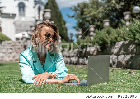 a young beautiful woman with blond curly hair in glasses and a blue dress sits on the grass in nature and uses a laptop, a young beautiful woman with blond curly hair in glasses and a blue dress sits on the grass in nature and uses a laptop, 98226999