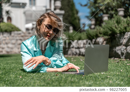 a young beautiful woman with blond curly hair in glasses and a blue dress sits on the grass in nature and uses a laptop, 98227005