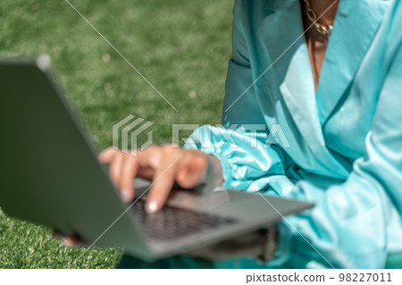 a young beautiful woman with blond curly hair in glasses and a blue dress sits on the grass in nature and uses a laptop, 98227011