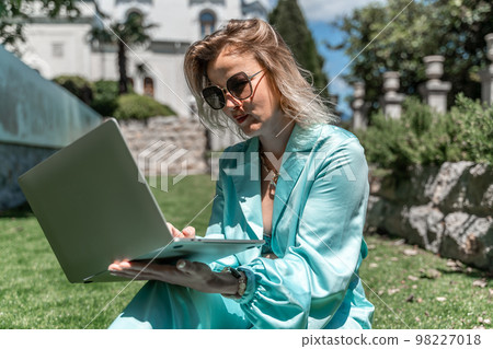 a young beautiful woman with blond curly hair in glasses and a blue dress sits on the grass in nature and uses a laptop, a young beautiful woman with blond curly hair in glasses and a blue dress sits on the grass in nature and uses a laptop, 98227018
