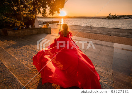A woman in a long red dress against the backdrop of sunrise, bright golden light of the sun's rays. The concept of femininity, harmony. 98227181