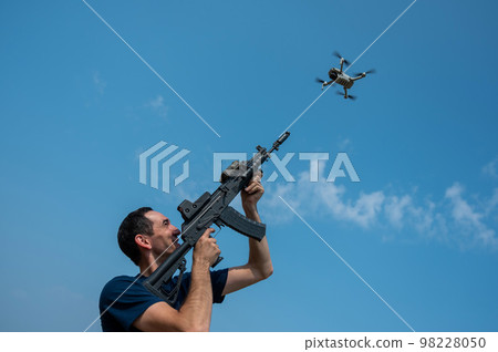 A man aims to shoot a rifle at a flying drone against a blue sky. A man aims to shoot a rifle at a flying drone against a blue sky. 98228050