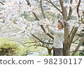 A smiling senior woman lifting a branch of cherry blossoms and looking at the flowers without an apron 98230117