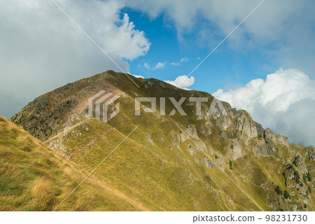 Sunny valley among majestic green cloudy mountain hills covered in green lush grass, and bushes. 98231730