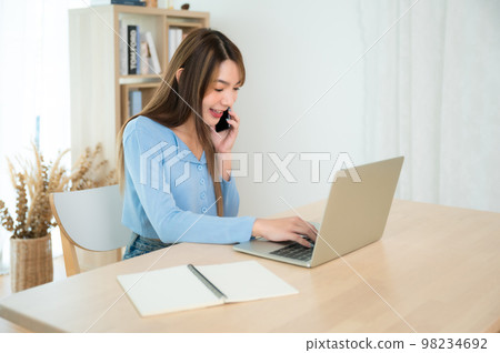 Young asian woman talking mobile smartphone while working on computer laptop Young asian woman talking mobile smartphone while working on computer laptop 98234692