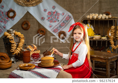 Little smiling girl in folk costume sprinklers pancakes with honey while celebrating Maslenitsa  98237614