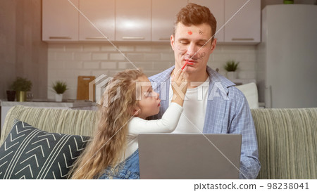 Preschooler girl paints lips of father working on laptop Preschooler girl paints lips of father working on laptop 98238041