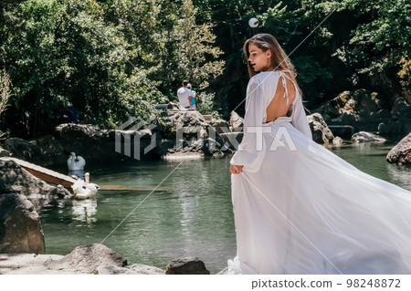 a beautiful woman in a long white dress looks into the distance at a beautiful lake with swans rear view a beautiful woman in a long white dress looks into the distance at a beautiful lake with swans rear view 98248872