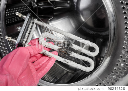 A man holds in his hand a burnt-out heating element of a washing machine against the background of a washing machine drum 98254828