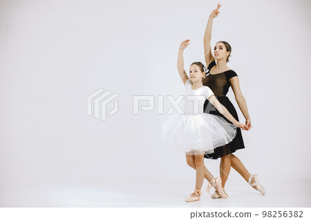 Little girl ballerina with her mother on white background. Brunette girl wearing white ballet suit and adult woman black dress. Two female dancer dancing together in a pointe shoes. Little girl ballerina with her mother on white background. Brunette girl wearing white ballet suit and adult woman black dress. Two female dancer dancing together in a pointe shoes. 98256382