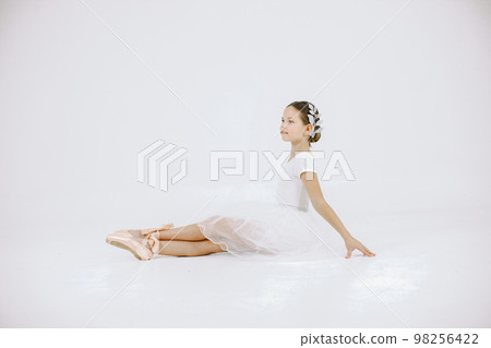 Little ballerina on white background. Brunette girl wearing white ballet suit. Girl sitting on a floor. 98256422