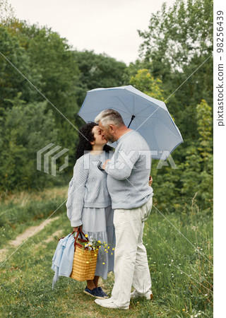Close up romantic couple standing in autumn park under umbrella in daytime. Man and woman wearing blue sweaters. Man and woman kissing. 98256439