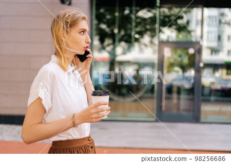 Side view of pretty elegance blonde female with cup of coffee to-go talking on smartphone. Cute young woman in casual white shirt speaking on mobile phone on summer day in city center street. Side view of pretty elegance blonde female with cup of coffee to-go talking on smartphone. Cute young woman in casual white shirt speaking on mobile phone on summer day in city center street. 98256686