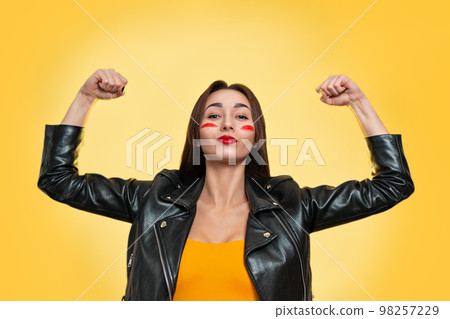 Feminism, girl power, and the concept of equal rights and independence for women. Caucasian young woman in a leather jacket shows her biceps. Isolate. Yellow background. Copy Feminism, girl power, and the concept of equal rights and independence for women. Caucasian young woman in a leather jacket shows her biceps. Isolate. Yellow background. Copy 98257229