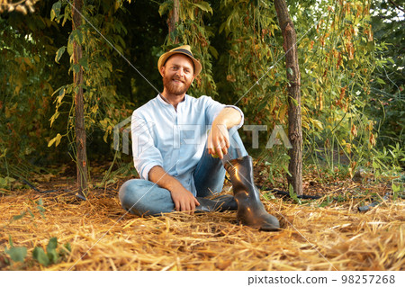 Summer organic gardening. Happy caucasian gardener wearing straw hat sits on the ground in greenhouse. The concept of local farming and Earth Day Summer organic gardening. Happy caucasian gardener wearing straw hat sits on the ground in greenhouse. The concept of local farming and Earth Day 98257268