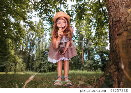 Cute little girl looks through a magnifying glass at the camera. Bottom view. Park in the background. The concept of scouting and children's education 98257288