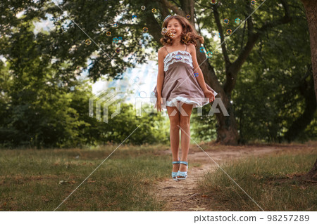 Happy childhood. Cute little girl in dress enjoying and jumping with soap bubbles at summer green park. Concept of Children's day and holidays 98257289