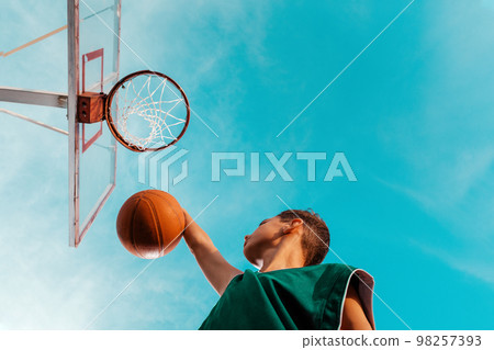 A young teenager in a green tracksuit is playing basketball, throwing a ball into the ring. Copy space. Bottom view. The sky in the background. Concept of sport and activity A young teenager in a green tracksuit is playing basketball, throwing a ball into the ring. Copy space. Bottom view. The sky in the background. Concept of sport and activity 98257393
