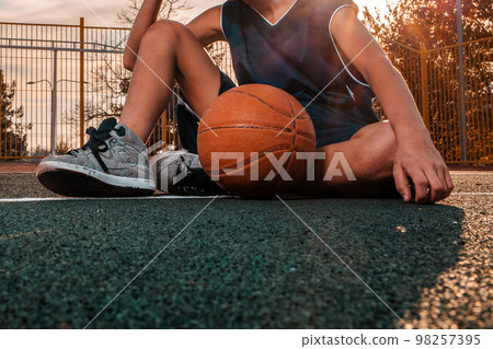 A young teenager in a blue tracksuit poses with a basketball ball sitting on the floor of a sports field. Close up. Sunset in the background. Concept of sport and activity A young teenager in a blue tracksuit poses with a basketball ball sitting on the floor of a sports field. Close up. Sunset in the background. Concept of sport and activity 98257395