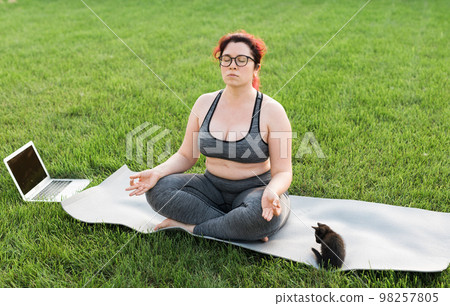 Plus size body positivity woman doing meditation on yoga mat with kitten on backyard - wellness and mental health concept 98257805