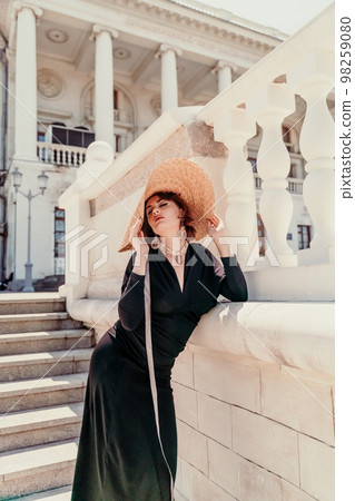 Fashion photo of a beautiful model in an elegant hat and black dress posing against the backdrop of a building with columns. 98259080
