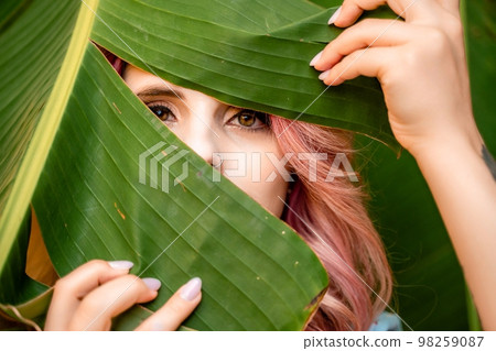 Woman portrait pink hair banana leaf. A beautiful young woman among the huge green leaves of a banana tree. 98259087