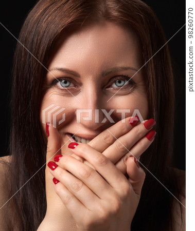 Cheerful charming woman laughing while covering mouth with her hands with red manicure on nails and gazing at camera. Studio portrait of Caucasian ethnicity 40-year-old brunette woman with gray eyes. 98260770