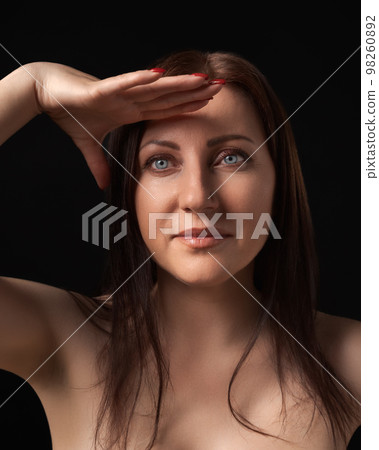 Pretty brunette woman gracefully raising hand to her forehead and looking at camera on black background. Studio portrait of beautiful and sexy woman with gray eyes and long hair. Sincere emotions 98260892