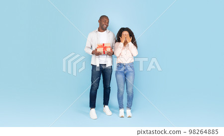 Greeting concept. Loving black man holding gift box, excited lady covering her eyes with hands and waiting for surprise 98264885