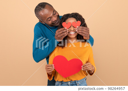 Valentine's holiday celebration concept. Loving black middle aged man covering wife's eyes with paper hearts Valentine's holiday celebration concept. Loving black middle aged man covering wife's eyes with paper hearts 98264957