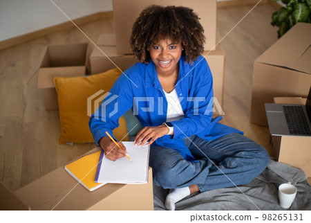 Black Woman Sitting Among Cardboard Boxes After Moving And Writing In Notepad Black Woman Sitting Among Cardboard Boxes After Moving And Writing In Notepad 98265131