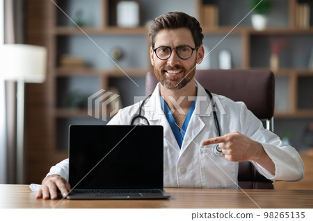Doctor pointing at blank laptop screen while sitting at desk in office Doctor pointing at blank laptop screen while sitting at desk in office 98265135