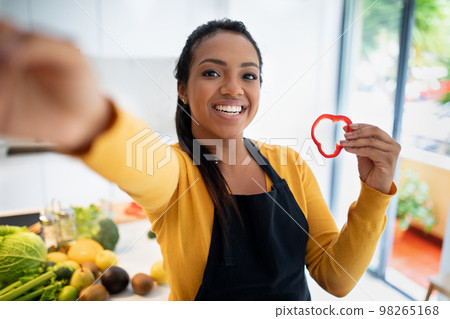 Smiling young african american female in apron with piece of pepper makes selfie with vegetables 98265168