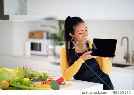 Happy young african american woman in apron show tablet with empty screen at table with fresh vegetables 98265175