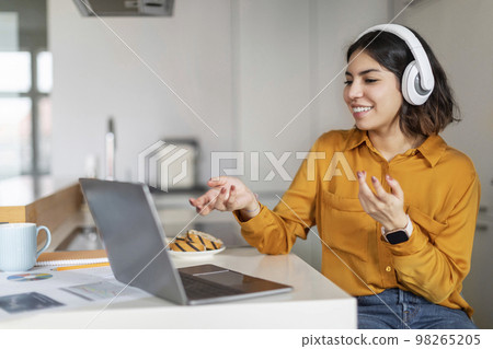 Video Call. Millennial Arab Woman In Headphones Teleconferencing On Laptop In Kitchen Video Call. Millennial Arab Woman In Headphones Teleconferencing On Laptop In Kitchen 98265205