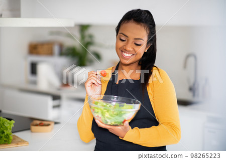 Smiling young african american female chef in apron eating organic vegetable salad, enjoy fresh dish 98265223