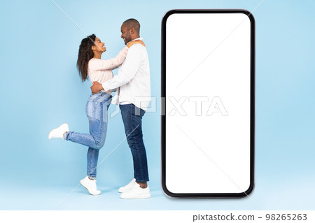 Loving black couple embracing and standing near large mobile phone with empty screen, full length, mockup Loving black couple embracing and standing near large mobile phone with empty screen, full length, mockup 98265263