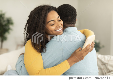 Cheerful young african american lady hugging man and chatting in smartphone, ordering goods in home interior 98265314