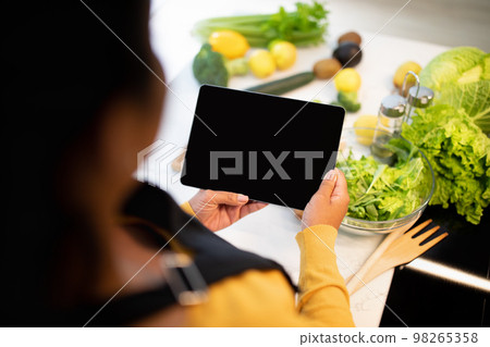 Young african american chef lady in apron cuts salad, use tablet with blank screen at table with vegetables Young african american chef lady in apron cuts salad, use tablet with blank screen at table with vegetables 98265358