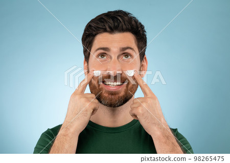 Bearded handsome man applying face cream on cheeks, standing on blue studio background, looking and smiling at camera Bearded handsome man applying face cream on cheeks, standing on blue studio background, looking and smiling at camera 98265475