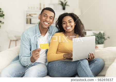 Cheerful young african american family use credit card, laptop and banking, check financial account, sit on sofa 98265637
