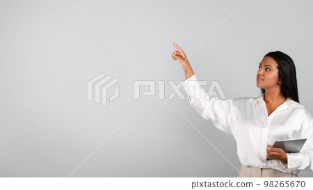 Smiling confident millennial black businesswoman with tablet controls virtual screen touches free space with finger Smiling confident millennial black businesswoman with tablet controls virtual screen touches free space with finger 98265670