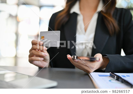 Businesswoman sits at her desk holding her phone and showing her credit card to camera 98266557